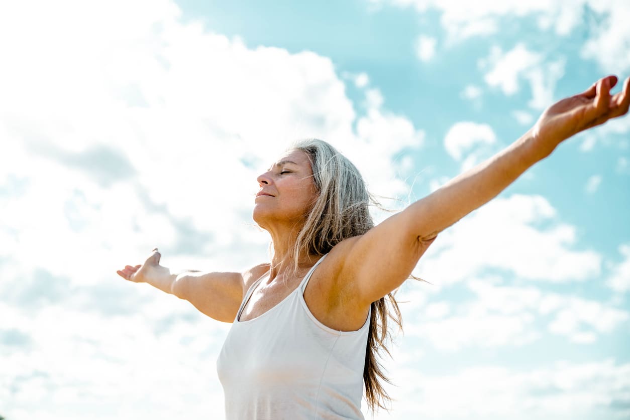 Joyful senior woman enjoying freedom standing with open arms and a happy smile looking up towards the sky - People and happiness concept